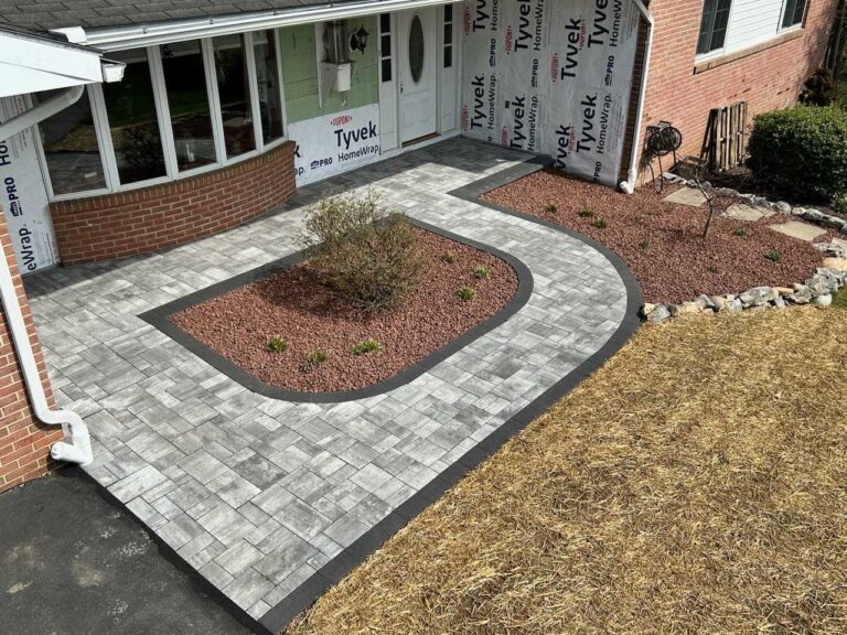 Newly installed gray paver patio with curved red stone planting bed around a shrub at the front entrance of a home