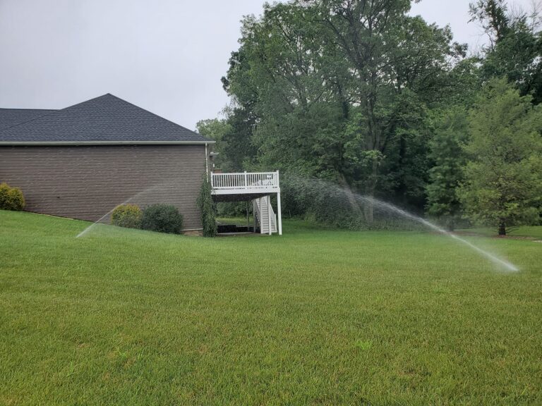 Lawn sprinkler system watering a large green lawn beside a residential home with elevated deck